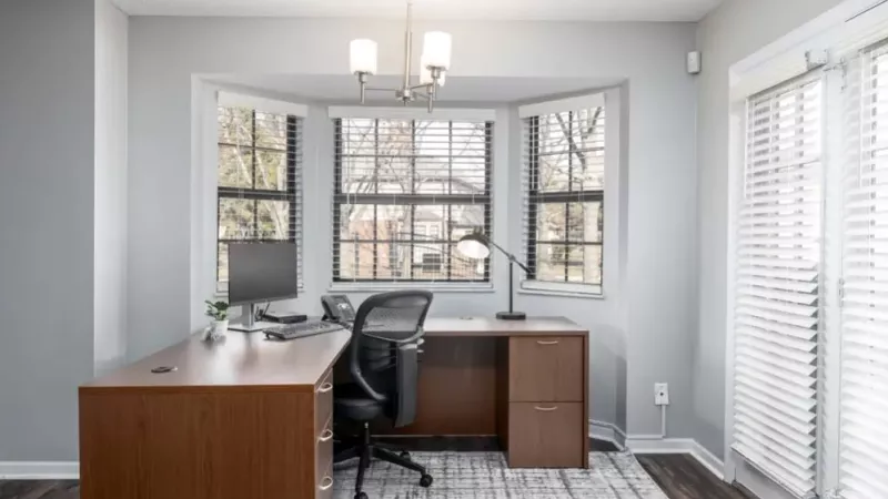 A stylish home office with a large wooden desk, ergonomic chair, and ample natural light from bay windows.