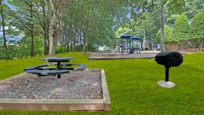 A picnic area with a circular table and a grill adjacent to a playground in a wooded setting.