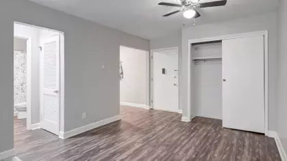 A well-lit room with wood flooring, a ceiling fan, and a built-in closet near a bathroom and entryway.