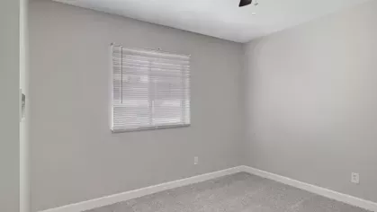 A cozy bedroom with gray carpeting, a ceiling fan, and a large window for natural light.