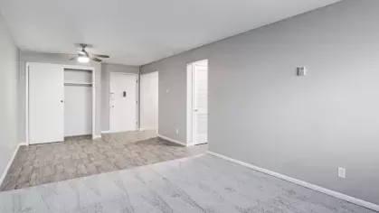 A spacious, unfurnished living room with light gray walls and carpeted flooring, featuring an open closet and ceiling fan.