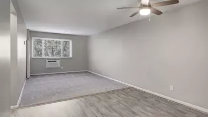 A long, well-lit living room with a large window and a ceiling fan, featuring both carpet and wood-style flooring.