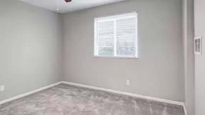 A carpeted bedroom with a large window and ceiling fan, featuring light gray walls and natural light.