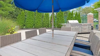 Outdoor dining area with a sleek BBQ grill station, wicker seating, and a large blue umbrella offering shade.