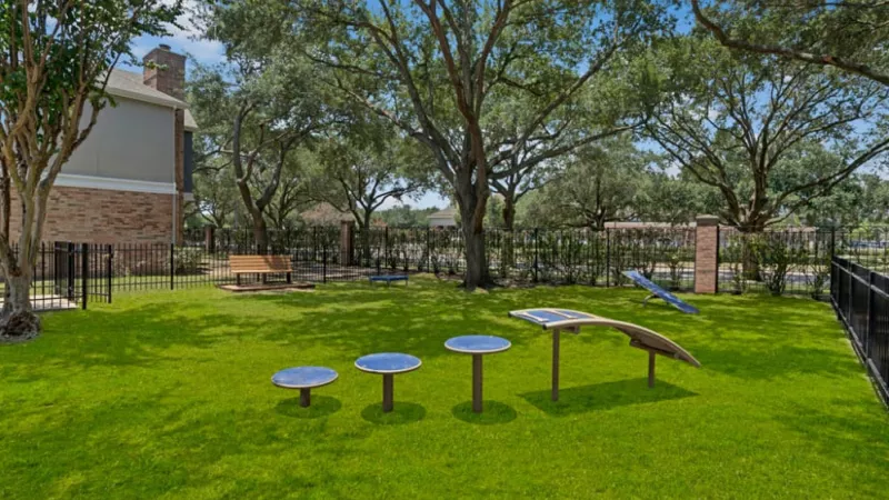 Fenced green space with exercise equipment and shaded benches under large trees.