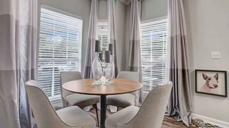 A charming dining nook with a round wooden table, upholstered chairs, and large windows with sheer curtains.