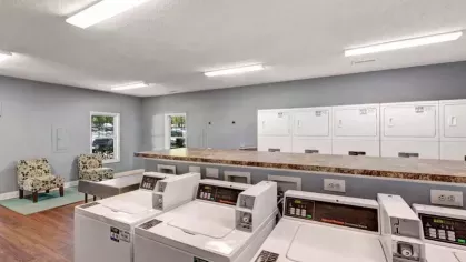 Spacious communal laundry room with multiple washing machines and dryers, a long folding counter, and seating with patterned chairs.