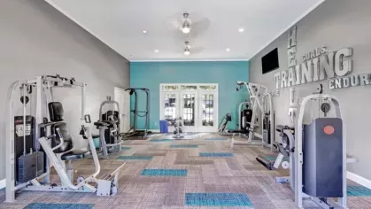 Modern fitness center with multiple workout machines, including weight stations and cardio equipment, featuring a blue and gray carpeted floor and motivational wall art.