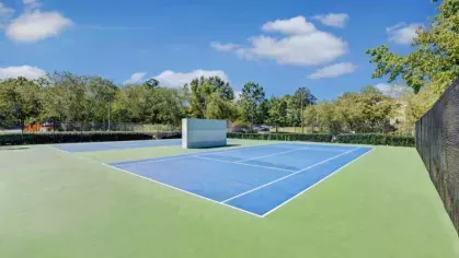 A vibrant blue tennis court sits under a clear blue sky, surrounded by lush greenery.