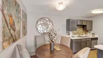 A cozy dining area with a round wooden table, four upholstered chairs, and decorative wall art, adjacent to the kitchen with gray cabinets and countertops.