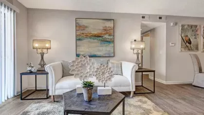 A stylish living room with a white sofa, two decorative table lamps, a large piece of abstract wall art, and a coffee table with a plant. Sliding glass doors let in natural light.