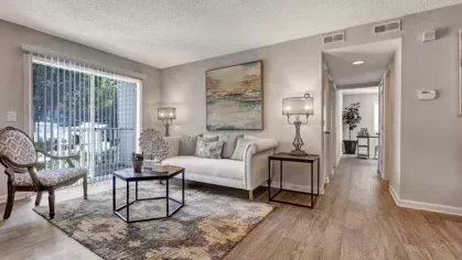 A bright living room with a white couch, a decorative coffee table, and sliding glass doors that lead to an outdoor area. A hallway leads to the bedrooms.