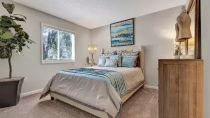 A stylish bedroom featuring a queen-size bed with light blue and gray bedding, a wooden dresser, and a large abstract painting above the headboard. A window allows natural light to brighten the space.