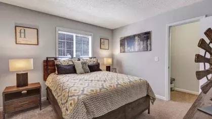 A cozy bedroom with a large bed, patterned bedding, and two nightstands with lamps. A framed wall print and window provide decor and natural light.