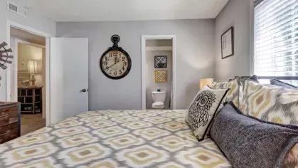 A different angle of the bedroom with patterned bedding, showing a door leading to an ensuite bathroom. A large clock is mounted on the wall.
