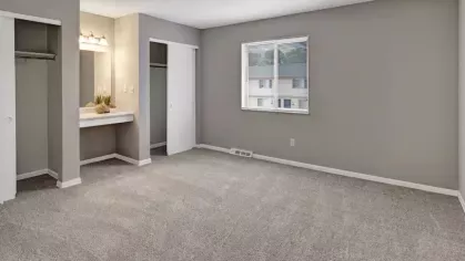 A spacious bedroom with neutral carpeting, a large window providing natural light, two open closets with sliding doors, and a built-in vanity area with a mirror and lighting.