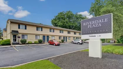 Image of the Riverdale Plaza Sign and Apartment Building: Riverdale Plaza Apartments sign in front of a two-story brick building with parking spaces in a neatly landscaped area.