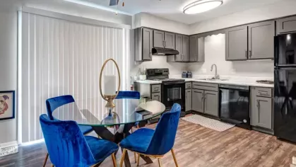 A dining area with a round glass table and blue velvet chairs, adjacent to a kitchen with gray cabinets and black appliances.