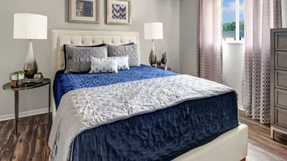 A bright bedroom with a tufted bed frame, blue and gray bedding, and matching side tables, featuring a window with natural light.