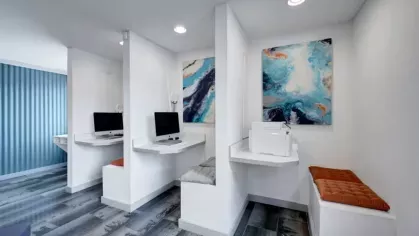 A small computer nook with white desks, colorful abstract art, and cushioned benches creates a cozy workspace.