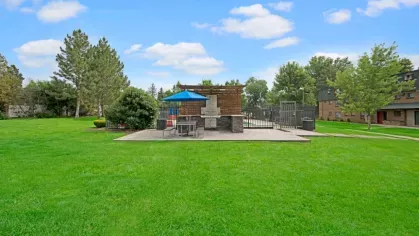 An outdoor grilling area with picnic tables and a blue umbrella surrounded by green grass.