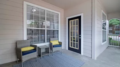 A welcoming patio area with two gray wicker chairs, yellow cushions, and a stylish rug outside a residential unit.