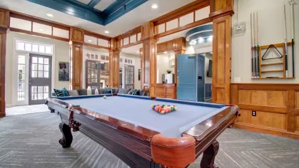 A recreational room featuring a billiards table with blue felt, pool cues mounted on the wall, and cozy seating.