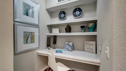 A small workspace nook with a built-in desk, shelves, decorative items, and framed wall art.