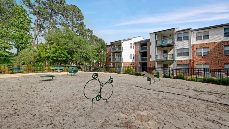 A sandy dog park with agility equipment, enclosed by fencing and overlooked by nearby residential buildings.