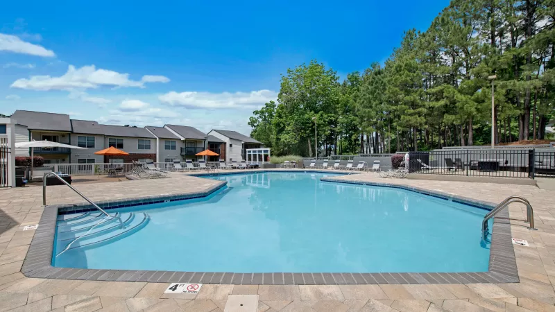 A wide-angle view of a swimming pool surrounded by lounge seating, with trees and apartment buildings in the background.