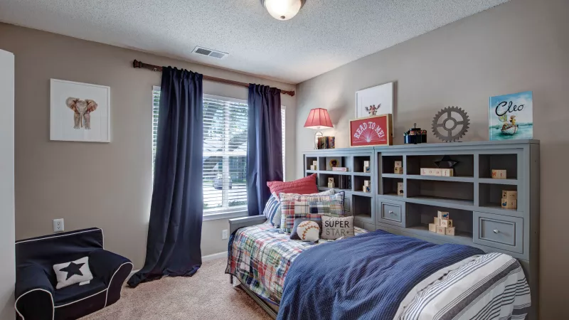 A cozy child’s bedroom featuring colorful bedding, a bookshelf filled with toys and books, and a bright window with navy curtains.