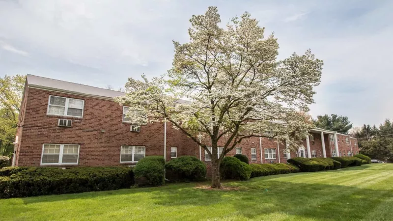 A charming red-brick apartment building surrounded by lush greenery, blooming trees, and well-maintained landscaping.