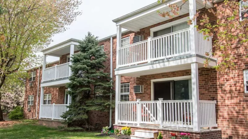 Red-brick apartments featuring white balconies with railing, surrounded by greenery, trees, and a beautifully landscaped yard.