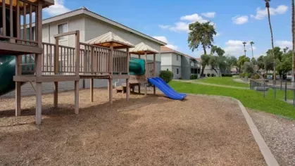 A fenced playground at 505 West Apartments, featuring a slide and climbing structures, surrounded by green lawns and shaded by mature trees.