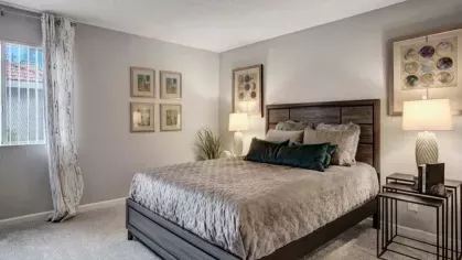 A spacious bedroom at 505 West Apartments featuring a modern wood-frame bed with decorative pillows, plush carpeting, and soft lighting, creating a warm and inviting retreat