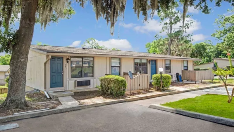 Exterior view of a one-story apartment building with light beige siding and blue doors, surrounded by neatly trimmed bushes and shaded by tall trees.