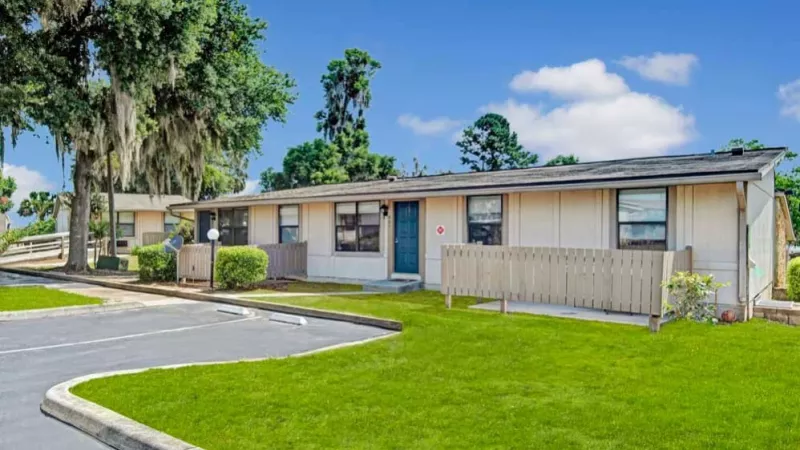 A cluster of one-story apartments with private patios and well-maintained green lawns, set against a clear blue sky.