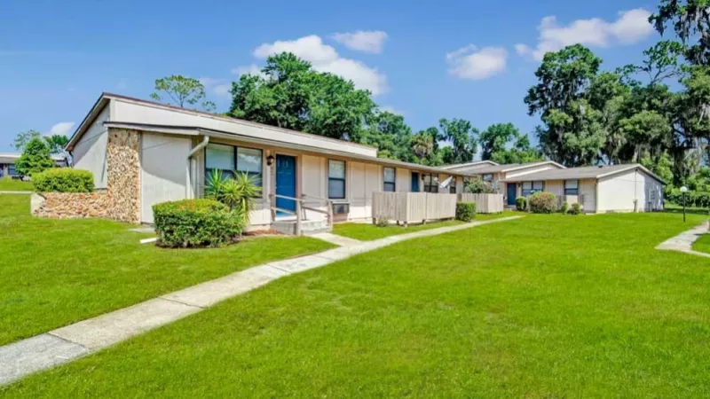 A row of single-story apartment homes with beige siding, blue doors, and wooden patios, set amidst expansive green lawns.