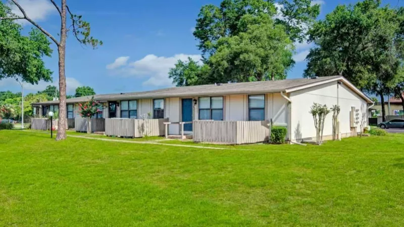 A row of single-story apartments with light-colored siding, fenced private patios, and surrounded by a lush green lawn under a clear blue sky.