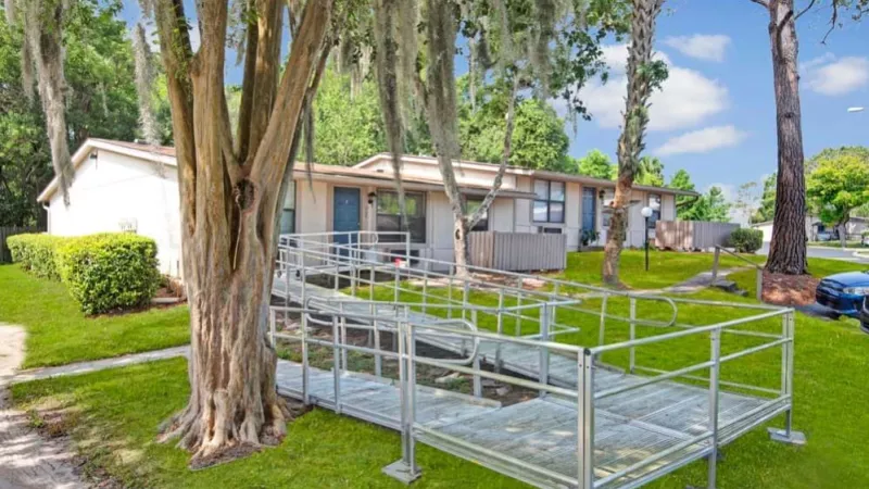 Apartments with accessible features, including a ramp and railings, surrounded by mature trees and a vibrant green setting.