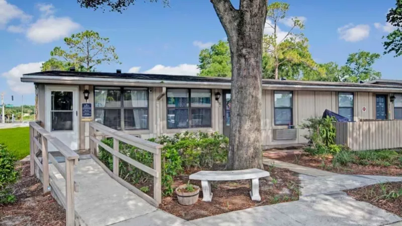 Exterior of Applewood Apartments with a ramp leading to the main office and a shaded bench in front of the building, surrounded by landscaping.