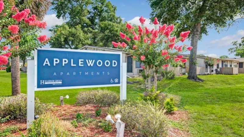 A close-up view of the Applewood Apartments sign framed by colorful flowers and trees, with residential buildings in the background.