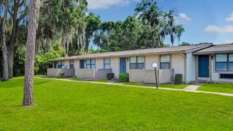 A row of single-story apartments with private patios, shaded by tall trees draped in Spanish moss, with a vibrant green lawn in the foreground.