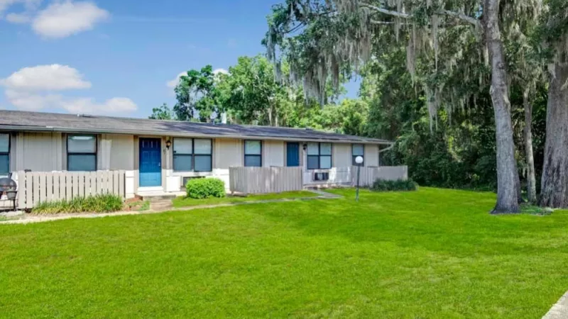 A row of single-story apartments with private entrances and light blue doors, set against a backdrop of greenery and a bright blue sky.