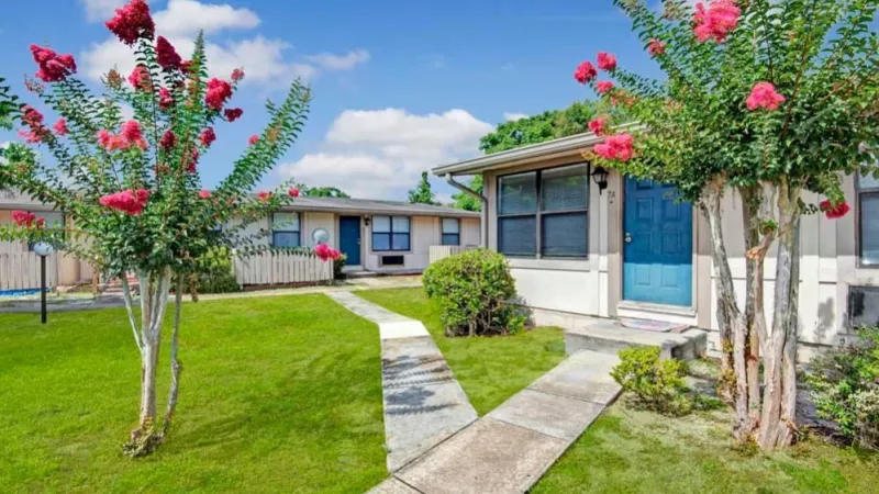 A landscaped pathway leading to single-story apartments with colorful crepe myrtle trees in full bloom and vibrant green grass surrounding the area.