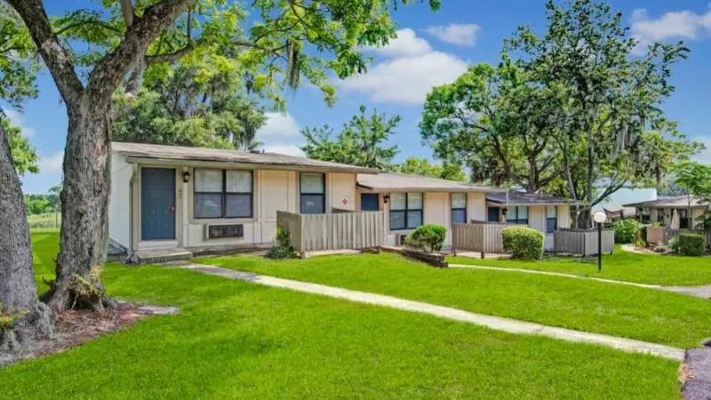 A serene view of single-story apartments with private patios and fenced areas, surrounded by mature trees and a manicured lawn.