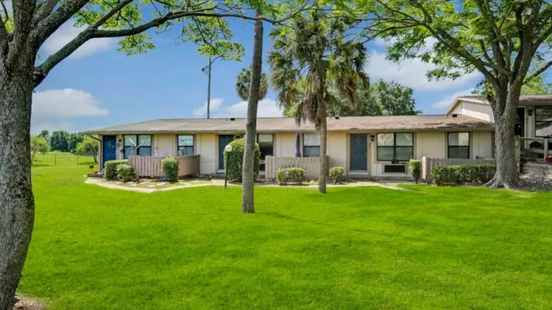 A group of single-story apartment homes framed by tall trees, with well-maintained green lawns and a walking path in the foreground.