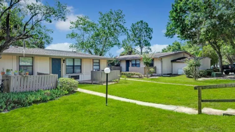 A quiet residential area with single-story apartments featuring private patios, shaded by large trees, and a grassy field nearby.