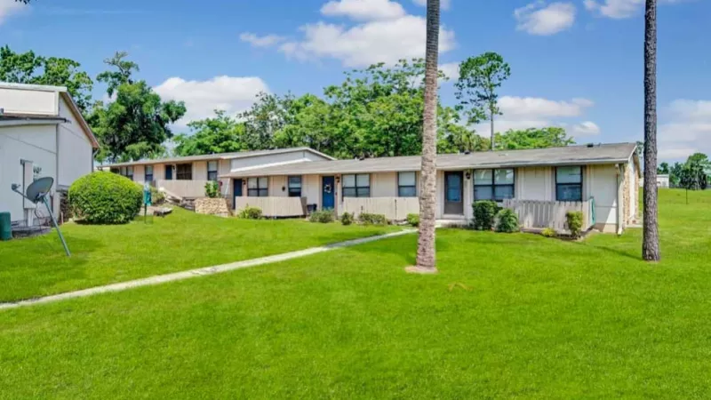 Single-story apartment units with private entrances, set against a backdrop of well-manicured lawns, palm trees, and a bright blue sky.
