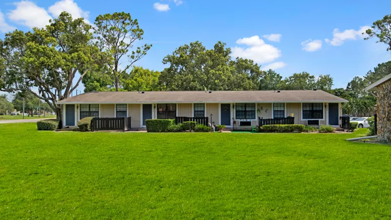 A row of cozy single-story apartment units with fenced porches and lush green lawns under a clear blue sky.